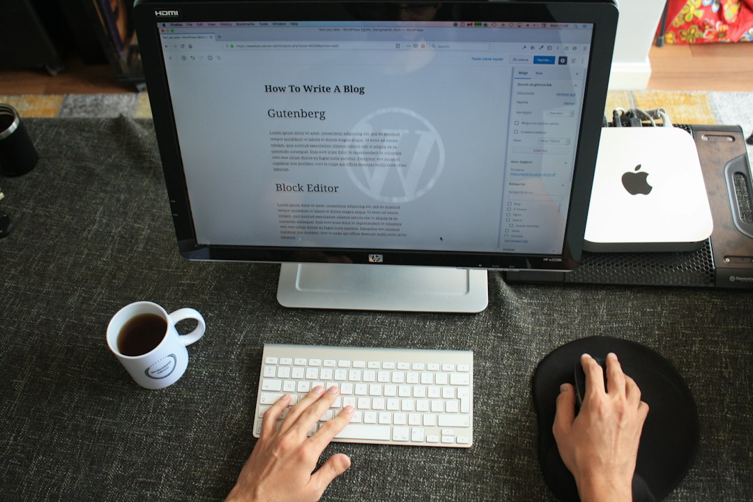 En person skriver på et hvidt tastatur ved et skrivebord og redigerer et blogindlæg om WordPress Hosting på en stor skærm. En kop kaffe, musemåtte og teknisk tilbehør er pænt arrangeret i nærheden.